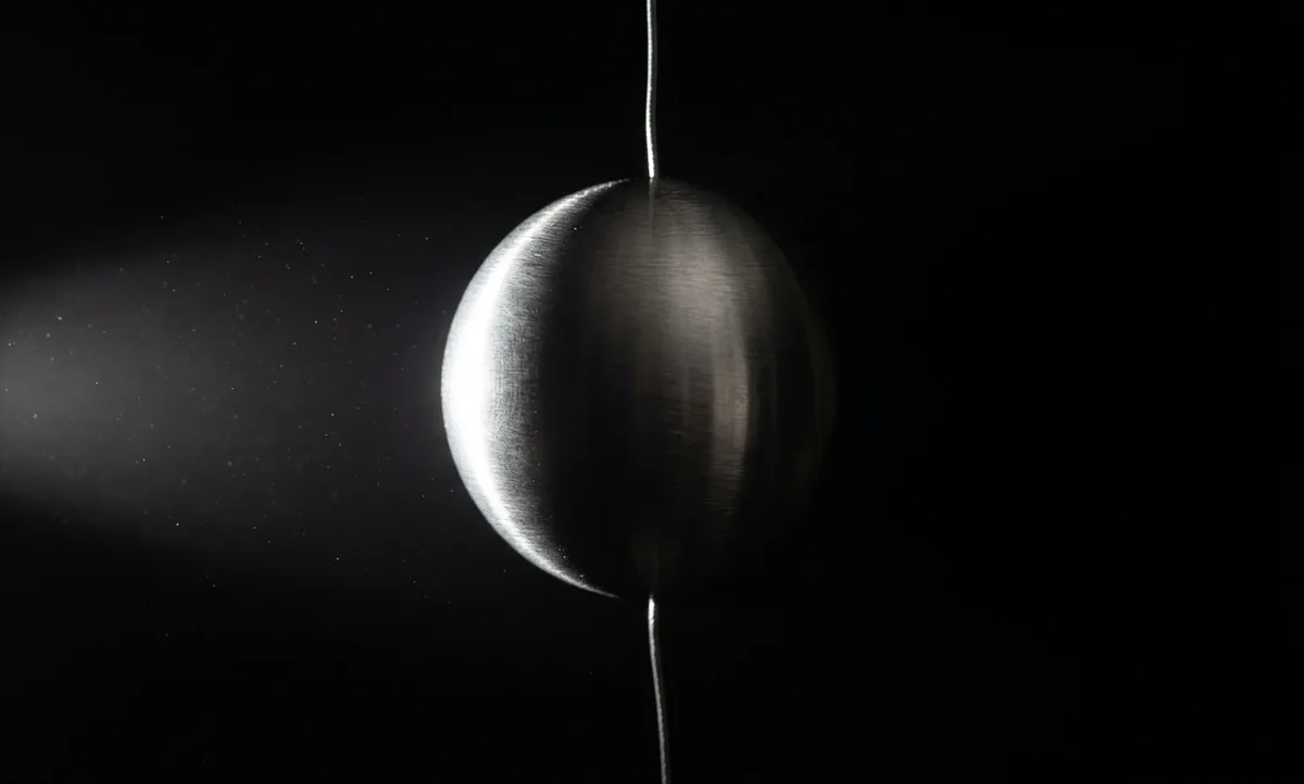Fotografia macro de uma esfera de metal escovado suspensa por um fio fino dentro de uma caixa preta, com o hemisfério esquerdo aceso por uma luz dura e o hemisfério direito quase apagado