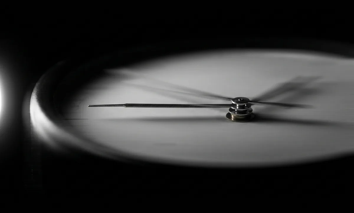 Fotografia macro do mostrador de um relógio analógico sem marca e sem números, com o ponteiro dos segundos nítido e parado enquanto o restante do mostrador aparece varrido por um borrão horizontal de movimento, fundo preto e luz lateral dura vinda da esquerda