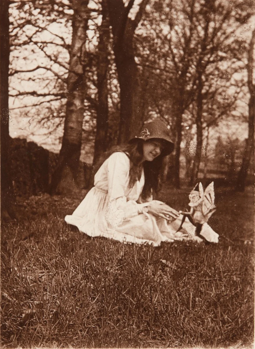 Elsie Wright de joelhos no gramado do quintal de Cottingley, em 1917, estendendo a mão para uma figura alada recortada em papel de cartão.