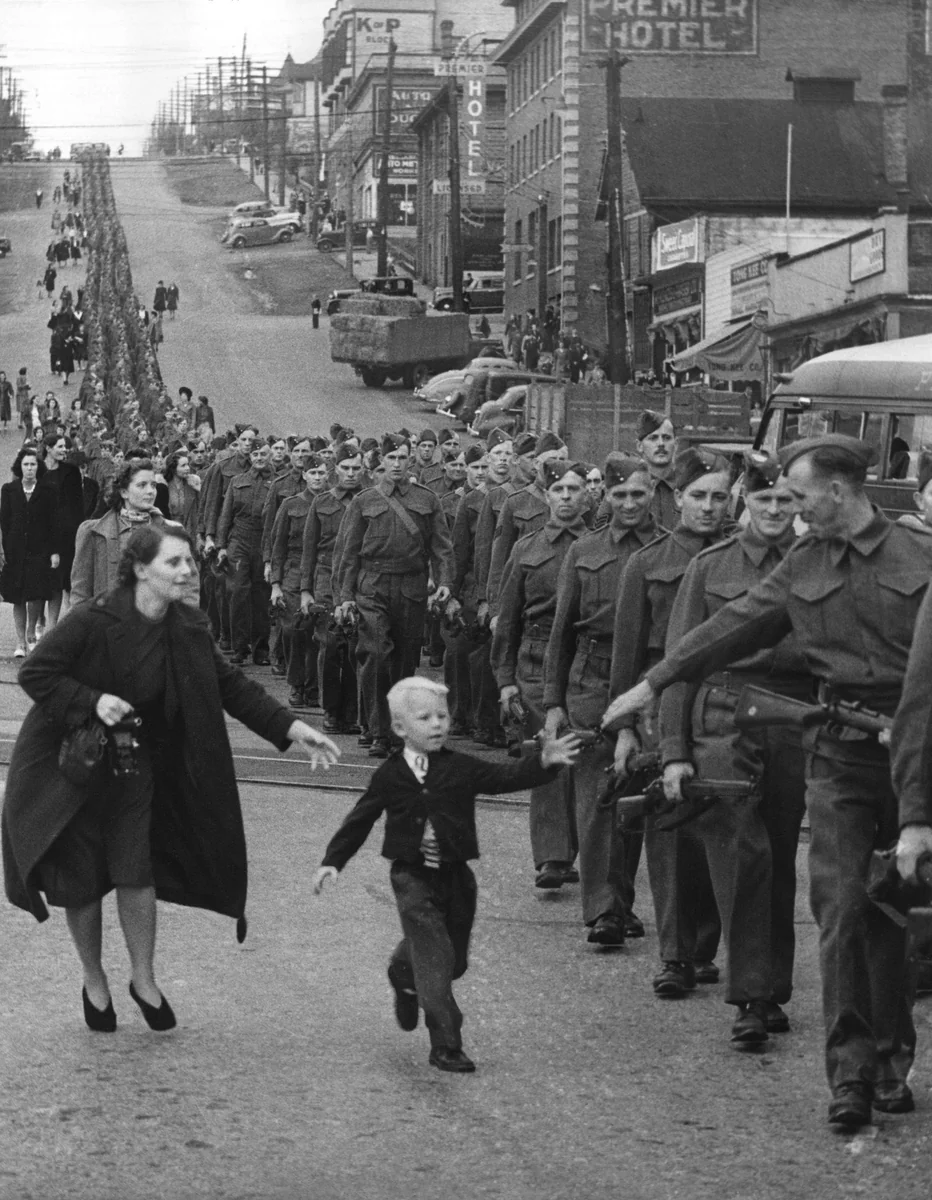 Menino de cinco anos correndo no meio da rua atrás do pai, soldado numa coluna do British Columbia Regiment em marcha, em New Westminster, 1940.
