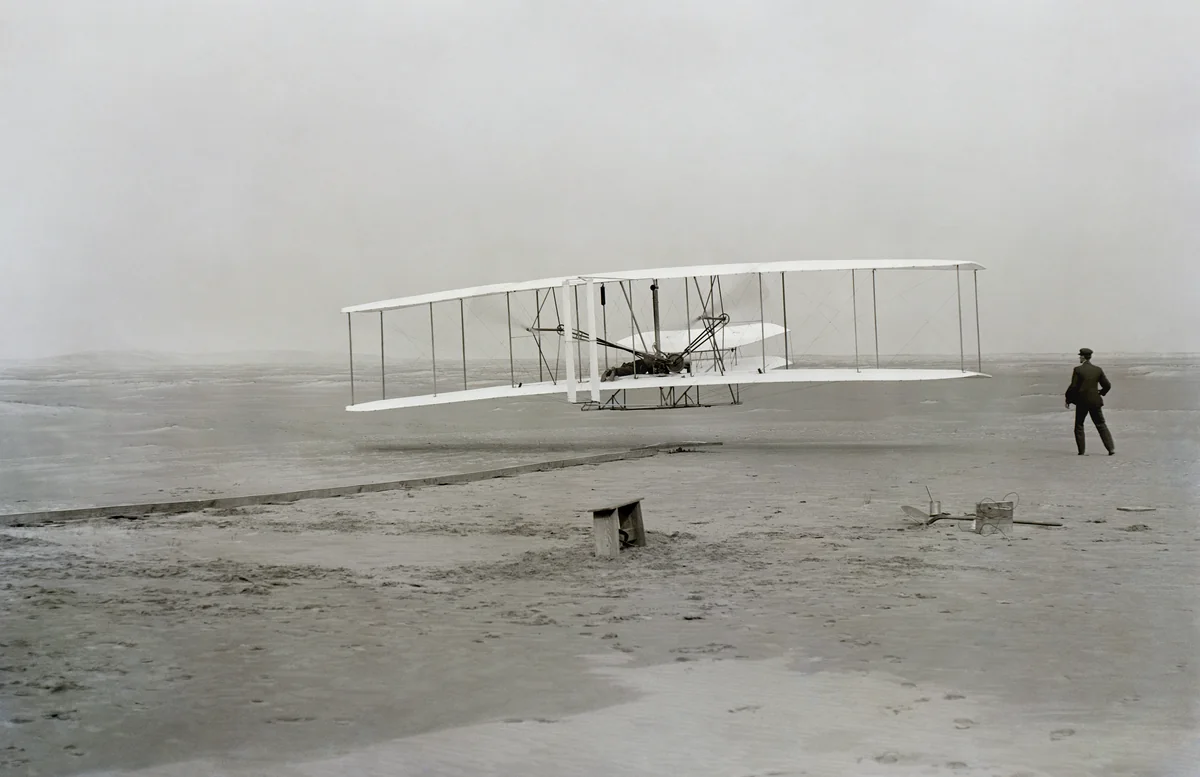 Wright Flyer a poucos palmos da areia em Kill Devil Hills, com Orville deitado sobre a asa inferior e Wilbur correndo ao lado, em 17 de dezembro de 1903.