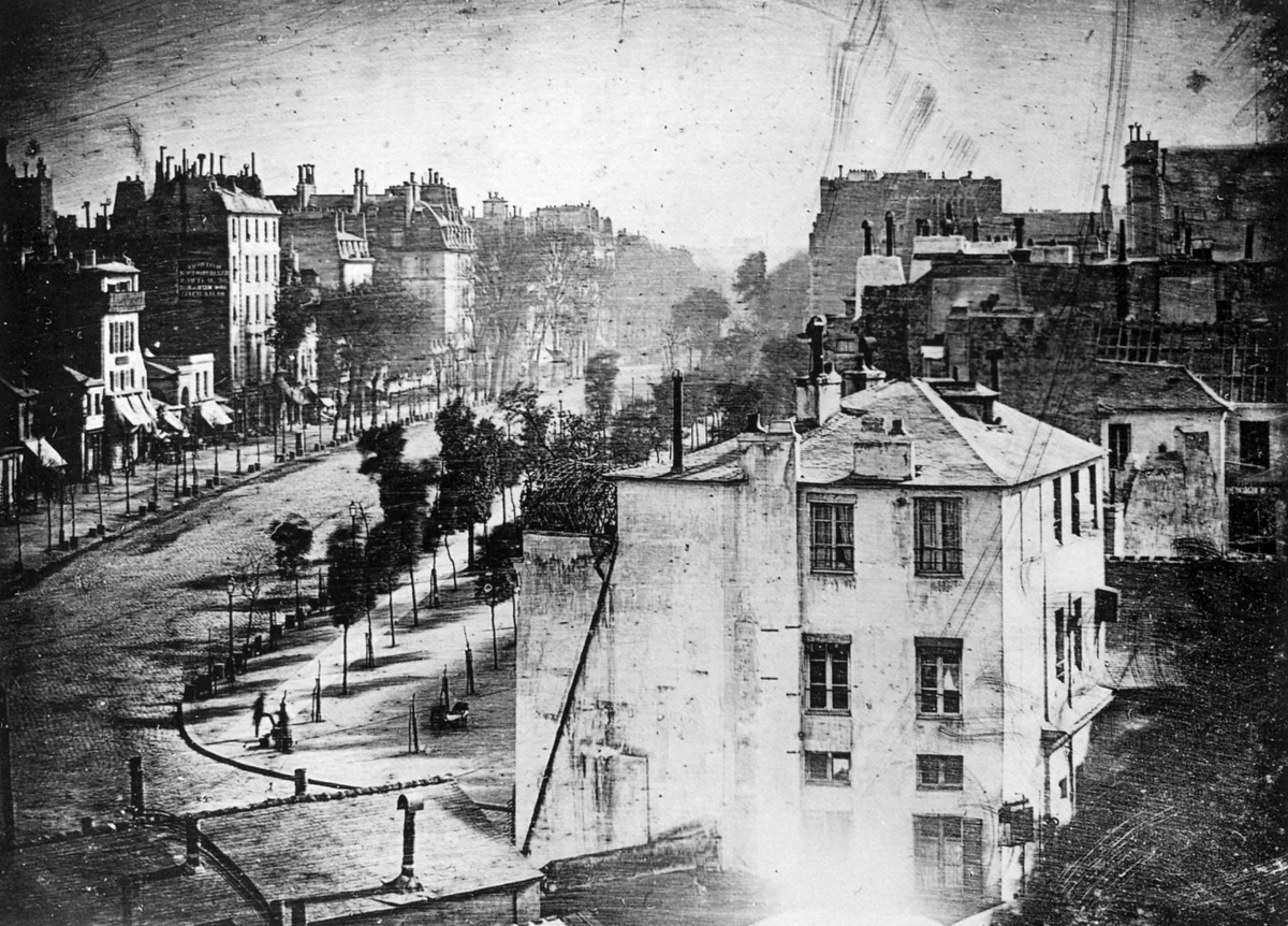 Vista do Boulevard du Temple em Paris feita por Louis Daguerre em 1838, com a calçada esvaziada pela longa exposição e dois homens imóveis no canto inferior esquerdo, um deles engraxando o sapato do outro.