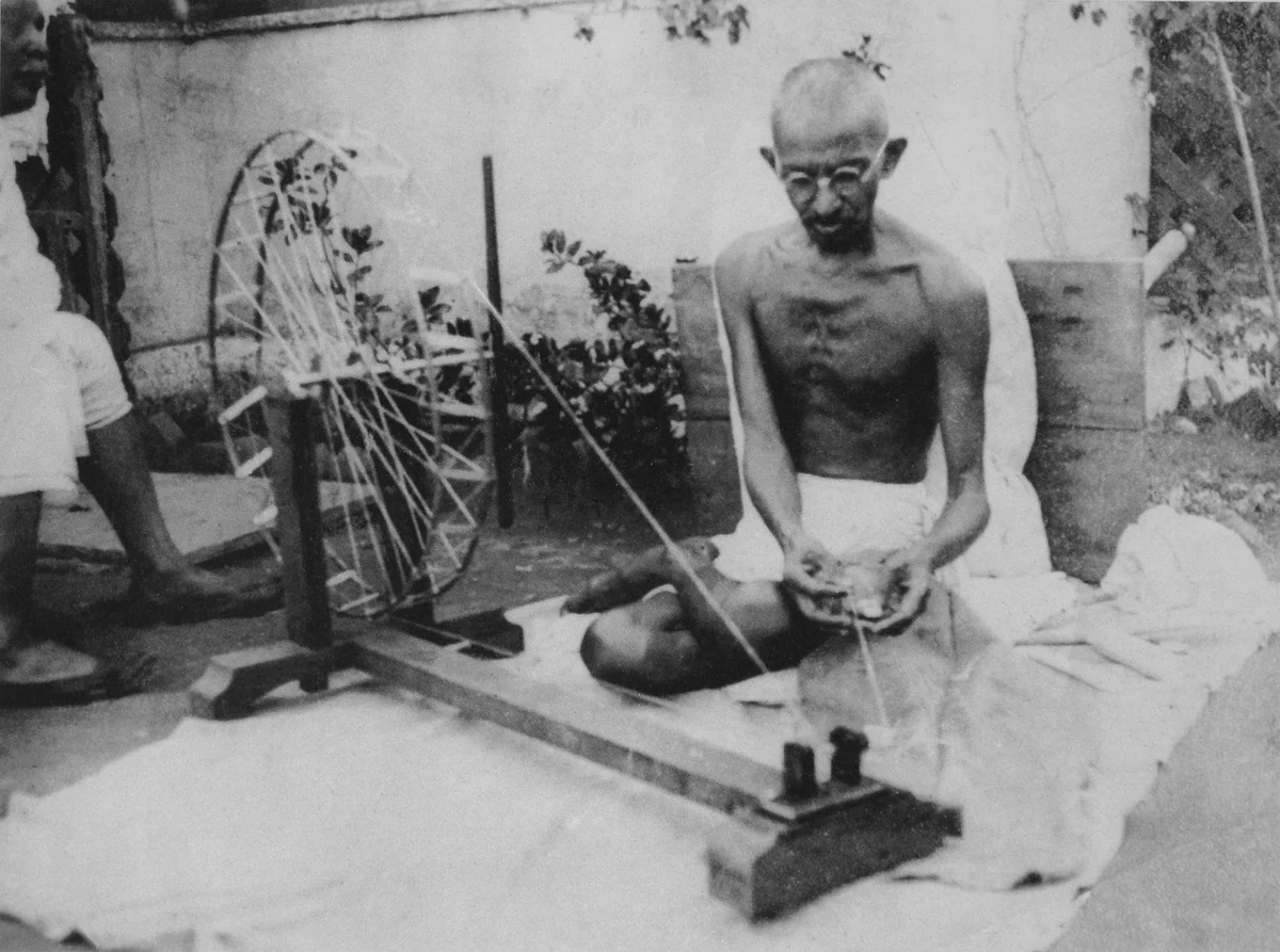 Gandhi sentado no chão, de pernas cruzadas, girando a roda da roca portátil de fiar algodão.