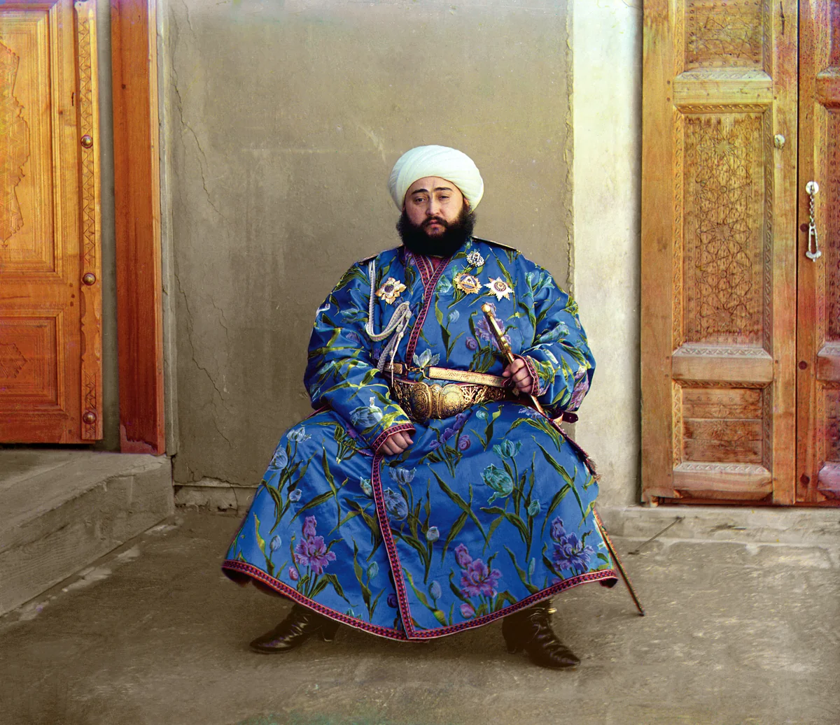 Alim Khan, emir de Bukhara, de pé com manto azul estampado, turbante branco, sabre e condecorações, retratado em cores em 1911.