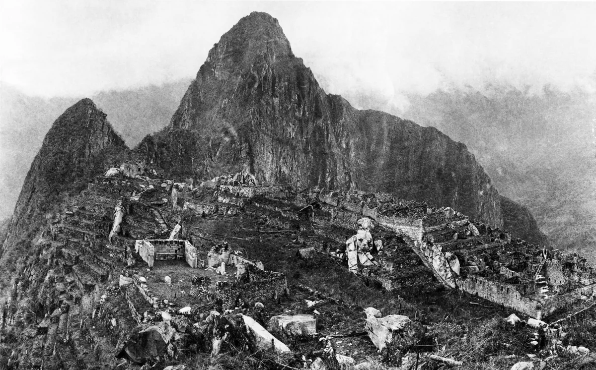 Vista geral de Machu Picchu em 1912, com terraços, praça e casas de pedra sem telhado e o pico Huayna Picchu ao fundo.