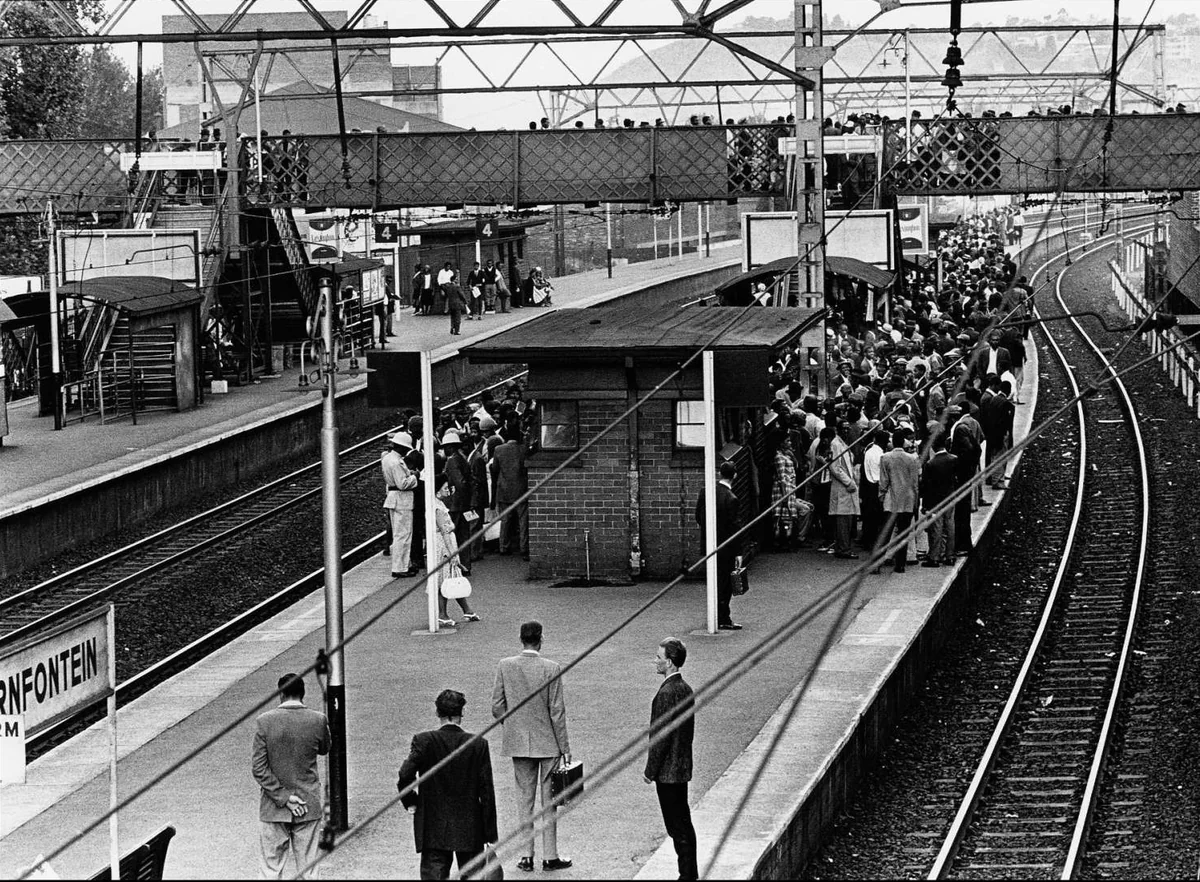 Plataforma da estação de Doornfontein, em Joanesburgo, com quatro homens brancos de terno em primeiro plano e a multidão de passageiros negros ao fundo, fotografada por Ernest Cole