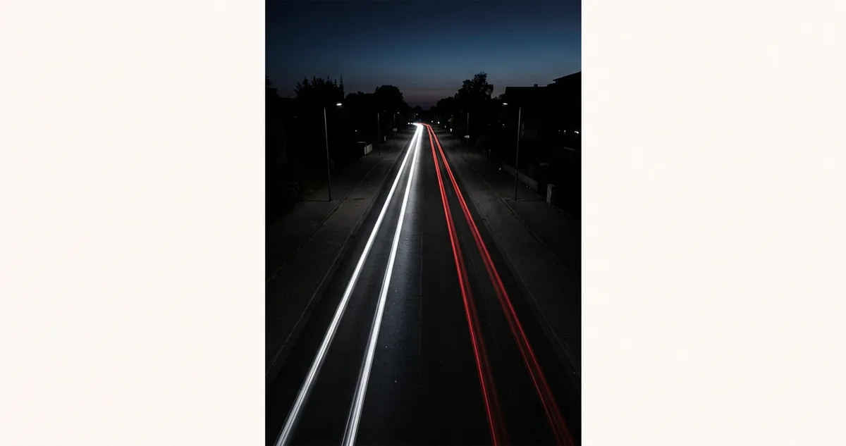 Avenida de mão dupla vista de cima à noite, com um traço de luz branco contínuo num sentido e traços vermelhos no outro, e nenhum carro visível na pista.