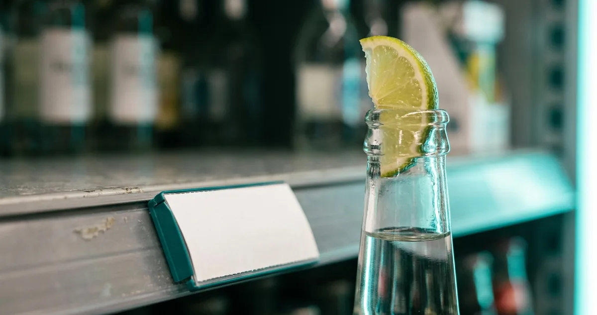 Garrafa transparente de Corona Extra com uma fatia de limão encaixada no gargalo, sobre o balcão de um bar.