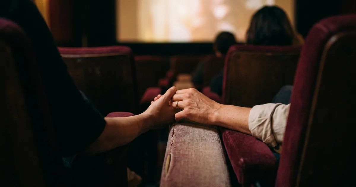 Duas mãos dadas entre poltronas de cinema, uma delas já escorregando pra fora